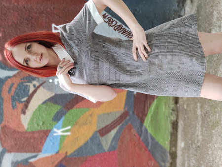 Pretty young redhead woman with a nice dress stands on the background of a wall with graffiti...