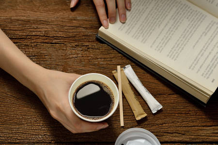 Woman holding paper cup of coffee and reading a book on wooden tableの写真素材