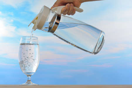 Fresh and clean drinking water being poured from jug into glass on sky backgroundの写真素材