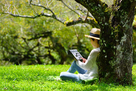 Happy middle aged woman sitting on green grass using tablet computer in the parkの写真素材