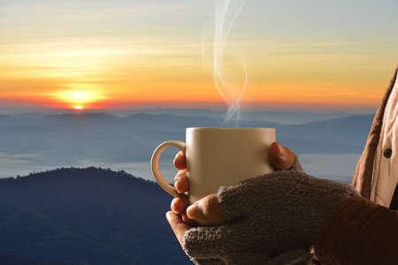 Hands in gloves holding coffee cup on mountain and sun background