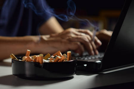 Man holding lit and smoking cigarette between fingers and working on a laptop computerの写真素材