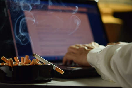 Man working on laptop computer with lit and smoking cigarette on an ashtray filled with cigarette buttsの写真素材