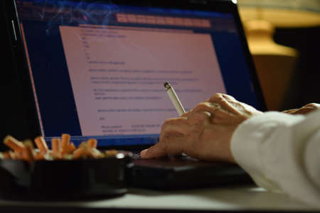 Man holding lit and smoking cigarette between fingers and working on a laptop computerの写真素材