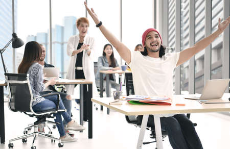 Young businessman raising both hands looking happy surrounded by group of business persons clapping in congratulationsの写真素材