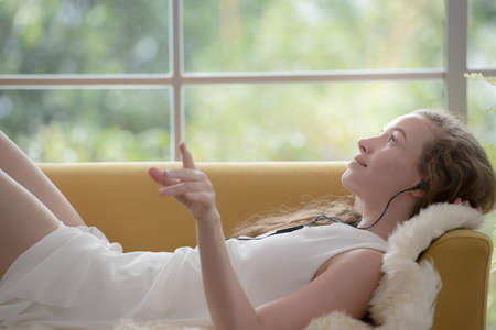 Beautiful young woman lying on a couch and listening to music on her cell phone looking relaxed and...