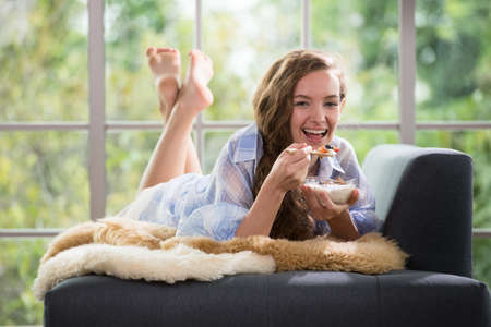Healthy young woman lying on a couch holding a bowl of yogurt looking relaxed and comfortableの写真素材