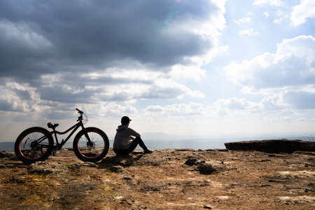 A person sitting down beside a bicycle on rocky mountain looking out at scenic natural view and beautiful blue skyの写真素材