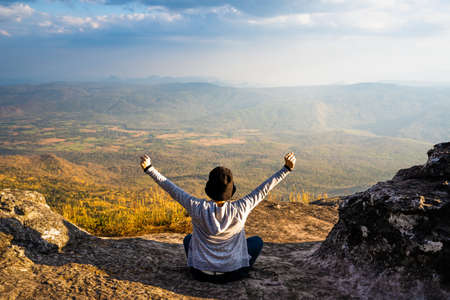 A woman sitting down with hands up on rocky mountain looking out at scenic natural view and beautiful blue skyの写真素材