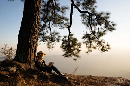 Woman sitting under pine tree reading and writing looking out at beaitiful natural view