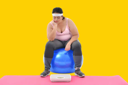 Overweight Asian woman sitting on gym ball looking boringly at the scaleの写真素材