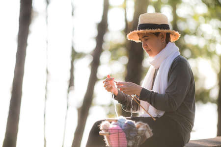 Middle aged Asian woman sitting on the bench and knitting for winter Christmas at beaitiful natural viewの写真素材