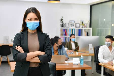 Asian woman wearing face mask smile and looking at camera working in new normal office and doing social distancing during corona virus covid-19 pandemicの写真素材