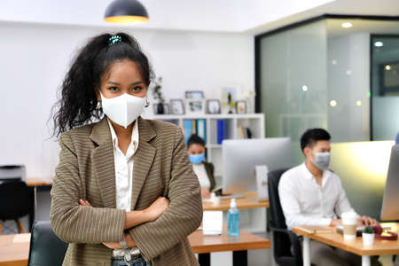 Asian woman wearing face mask smile and looking at camera working in new normal office and doing...