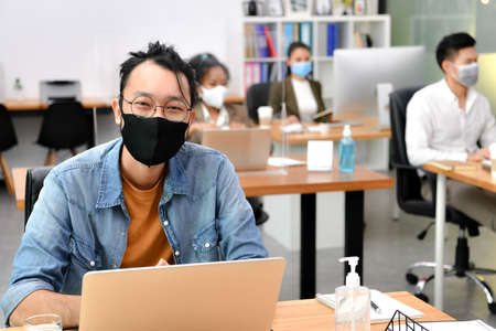 Asian man wearing face mask smile and looking at camera working in new normal office and doing social distancing during covid-19 pandemicの写真素材