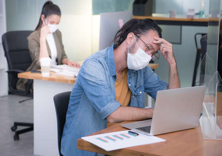 Asian man office worker wearing face mask working in new normal office and doing social distancing during corona virus covid-19 pandemicの写真素材