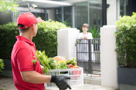 Asian delivery man in red uniform delivering  groceries box of food, fruit, vegetable and drink to woman recipientの写真素材