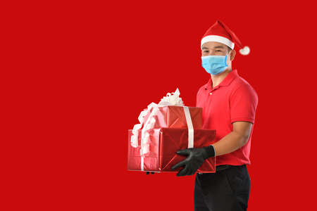 Happy young Asian delivery man in red uniform, medical face mask, protective gloves, christmas hat carry boxes of presents in hands on red background during COVID-19 outbreak and christmas festivitiesの写真素材