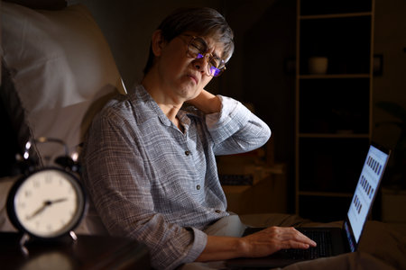 Senior Asian woman with sore and muscle pain from laptop use in bedの写真素材