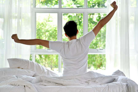 Asian man waking up in the morning sitting on bed and stretching