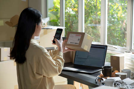Asian female small business owner using mobile app on smartphone checking parcel box. Warehouse...