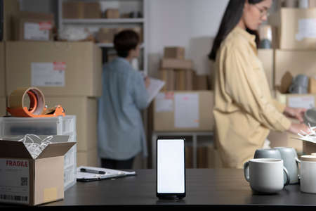 Smartphone with blank white screen standing on the desktop in the warehouses. Asian women online...