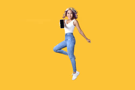 Full length portrait of young Asian woman holding smartphone jumping on yellow background. Cheerful young female jumping up and showing mobile phone with empty screen, in studioの写真素材