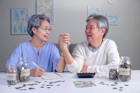 Savings concept. Asian old elderly senior couple grandparents husband and wife saving money, economizing pension, putting coin into money box, thinking over pension planの写真素材