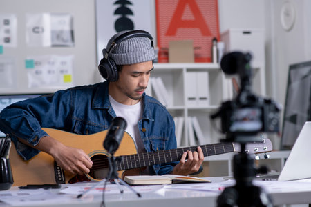Young Asian male graphic designer blogger influencer playing guitar while shooting education tutorial vlog training filming video course for social media at studio.の写真素材