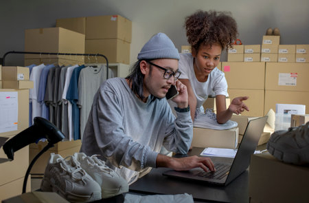 Mixed race couple of Asian man and African-American woman online seller confirming orders from customer on phone and laptop computer.の写真素材