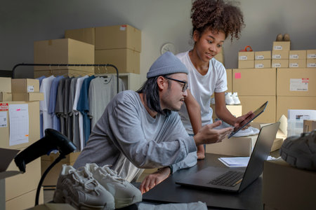 Mixed race couple of Asian man and African-American woman online seller confirming orders from customer on laptop and tablet computer.の写真素材