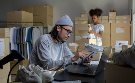 Mixed race couple of Asian man and African-American woman online seller confirming orders from customer on laptop and tablet computer.の写真素材