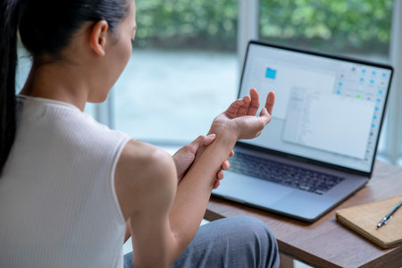 Asian woman is sitting with her laptop and her wrist aches while working at home office. Office syndrome hand pain by occupational disease.の写真素材
