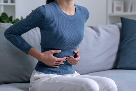 Young woman suffering from strong abdominal pain while sitting on sofa at home. Sick female having a stomach ache.の写真素材