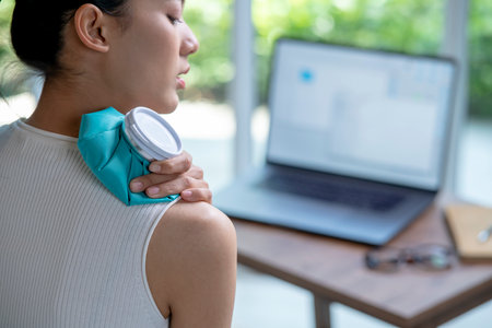 Asian woman putting a hot water bag on her shoulder pain while using laptop to work from home.の写真素材