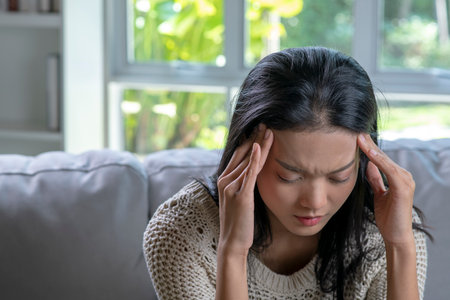 Headache. Young Asian woman is sitting on a sofa with her eyes closed, touching her head while suffering from a migraine.の写真素材