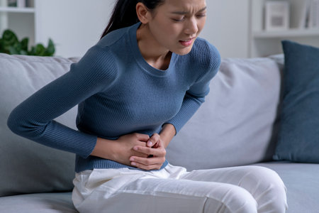 Young Asian woman suffering from strong abdominal pain while sitting on sofa at home. Sick woman having a stomach ache.の写真素材