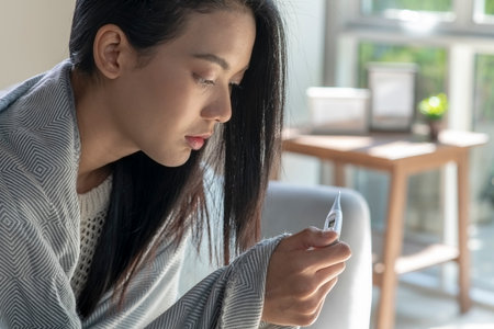 Sick young Asian woman at home on the sofa, she is covering with a blanket and taking temperature with a thermometerの写真素材