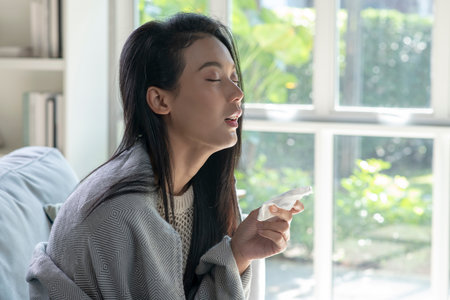 Ill young Asian woman covered with blanket blowing running nose got fever caught cold sneezing in tissue sit on sofa, sick allergic girl having allergy symptoms coughing at homeの写真素材
