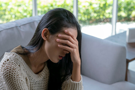 Headache. Young Asian woman is sitting on a sofa with her eyes closed, touching her head while suffering from a migraine.の写真素材