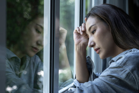Portrait of sad and worried Asian woman looking through the window at homeの写真素材