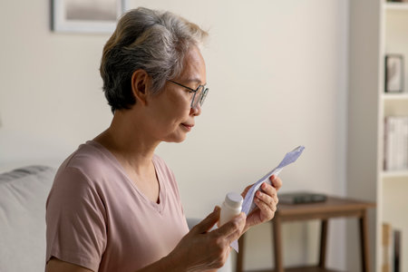 Asian senior woman sitting on sofa in her living room reading the information sheet of her prescribed medicineの写真素材