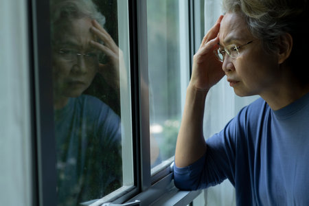 Portrait of sad and worried Asian senior woman looking through the window at homeの写真素材
