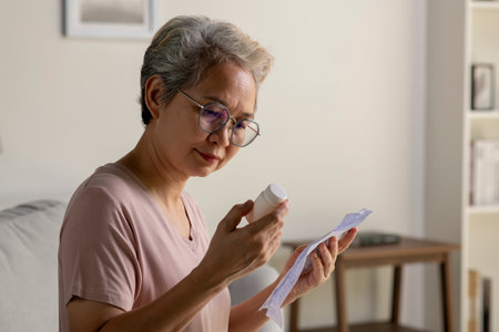 Asian senior woman sitting on sofa in her living room reading the information sheet of her prescribed medicineの写真素材