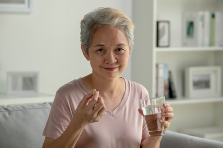 Asian senior woman taking medicine at home, smiling and looking at cameraの写真素材
