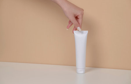 Woman hand is picking up the blank cosmetic packaging mockup: white tube, bottle on beige background.の写真素材
