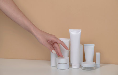 Woman hand is picking up the blank cosmetic packaging mockup: white tube, spray, bottle on beige background.の写真素材