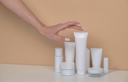 Woman hand is picking up the blank cosmetic packaging mockup: white tube, spray, bottle on beige background.の写真素材