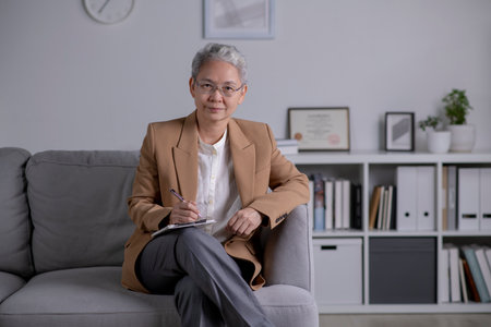Image of professional Asian female psychologist writing in clipboard, looking and smiling at camera, working in modern office. Psychotherapy services, mental health professional conceptの写真素材