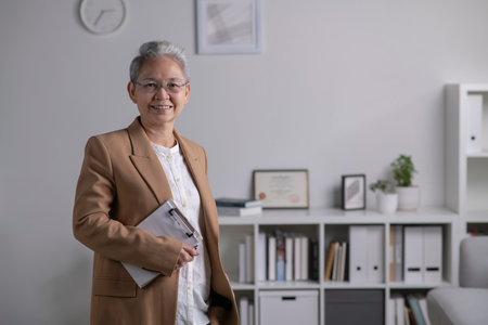 Image of professional Asian female psychologist standing, looking and smiling at camera, working in modern office. Psychotherapy services, mental health professional conceptの写真素材